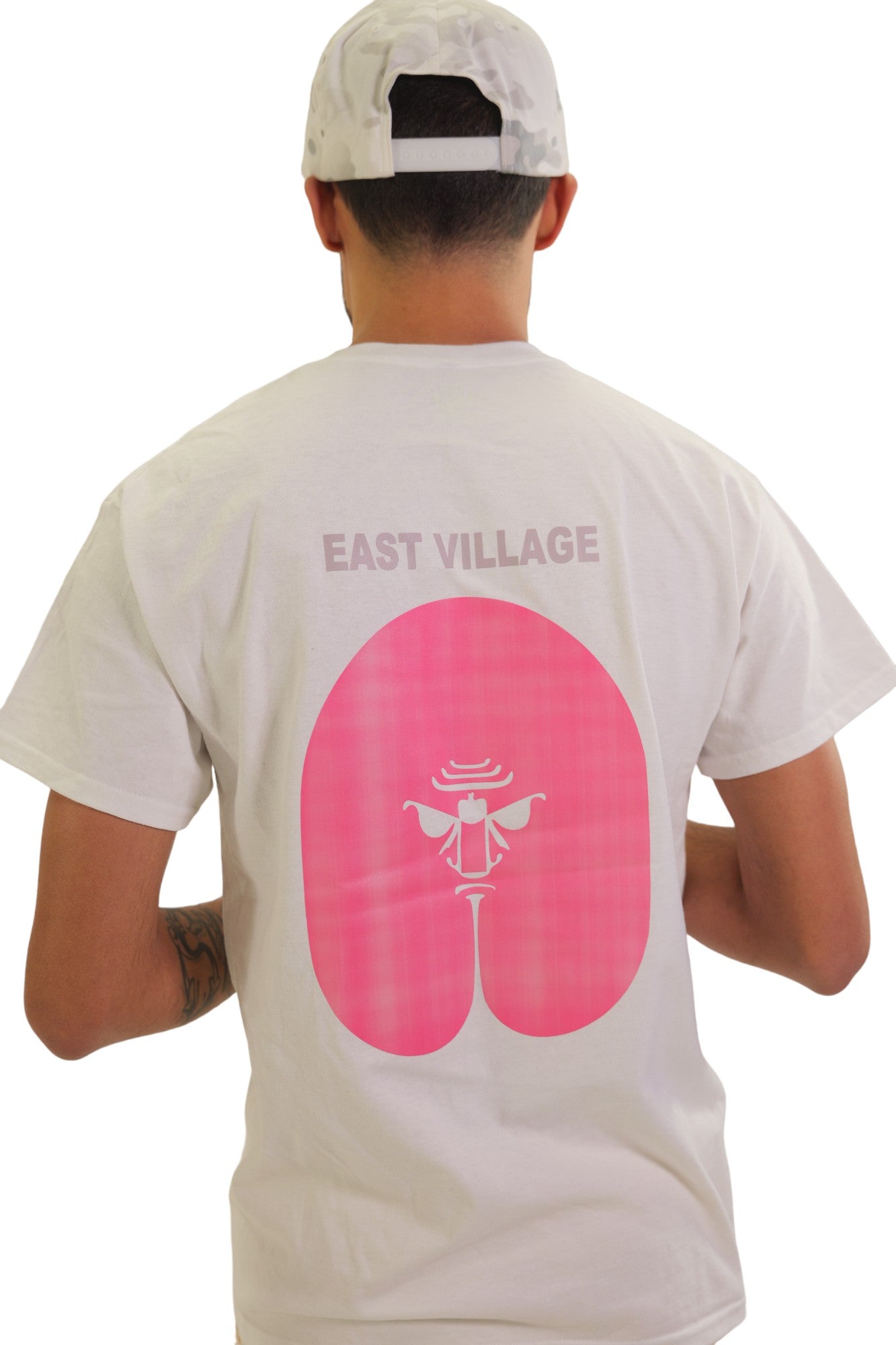 Back view of a model in a white t-shirt. The back has a large pink graphic of a butt with an angry face drawn in the center, and the words 'EAST VILLAGE' in grey above it