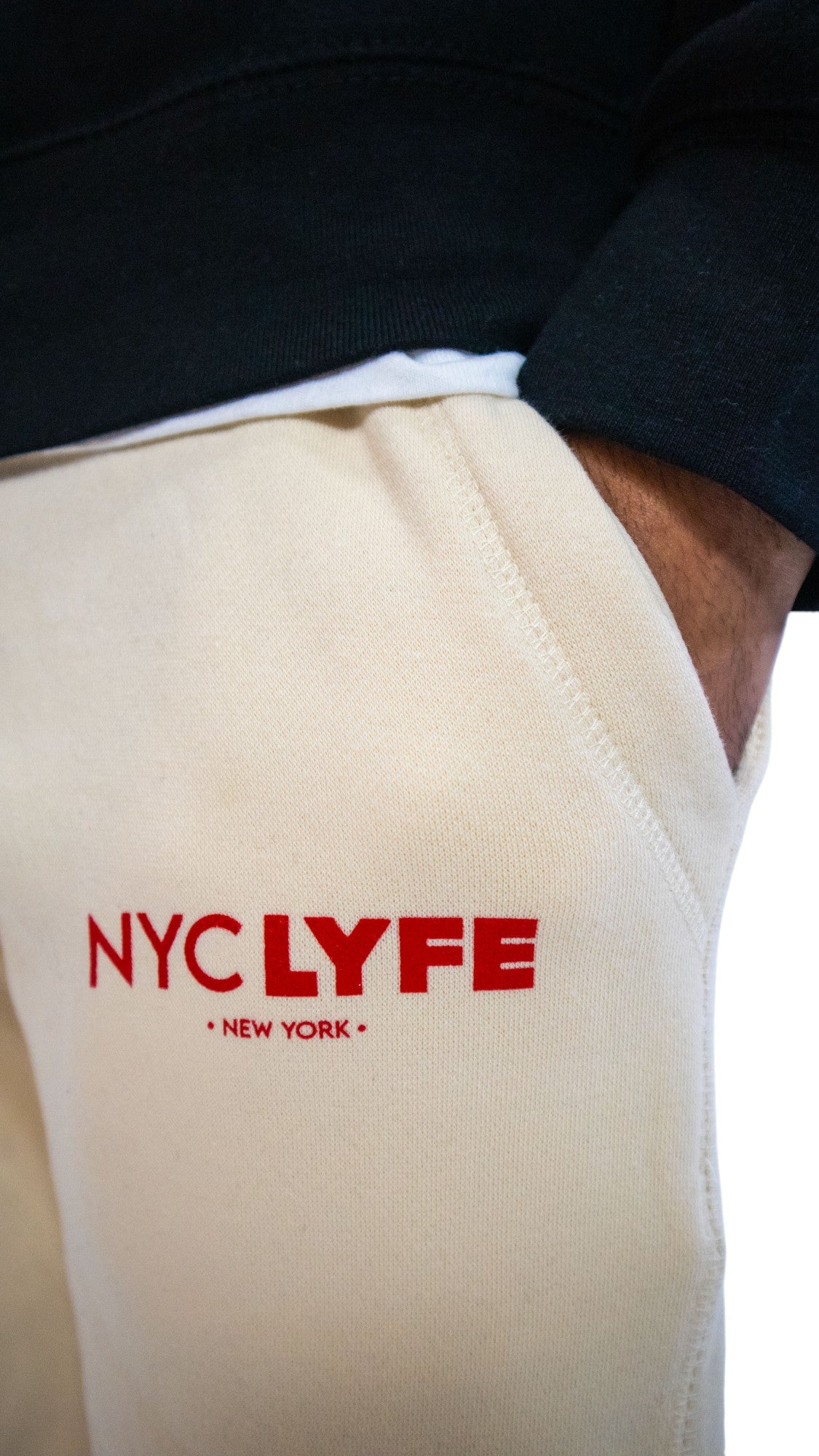 A close-up of off-white fleece shorts with a raw, cut-off hem. The 'NYC LYFE • NEW YORK •' logo is printed in red on the left thigh.