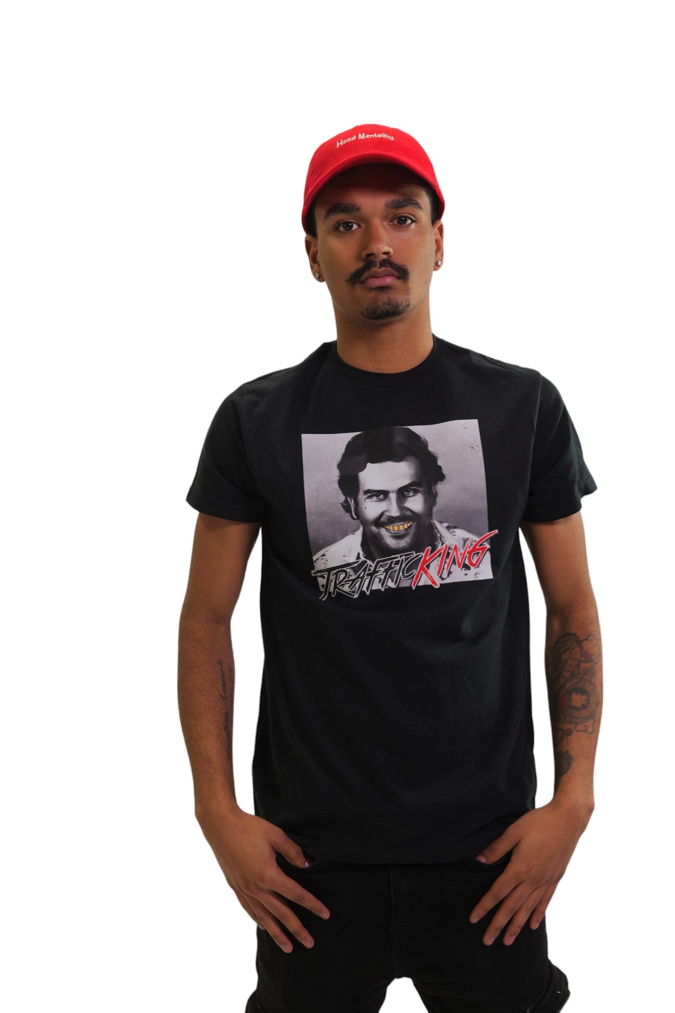 A young man with a mustache poses in a black short-sleeve t-shirt and a red baseball cap against a white background. The t-shirt has a graphic of Pablo Escobar with his teeth colored gold and bold red text over it that says 'Trafficking King'. The red cap has 'Hustle Mentality' embroidered in white.