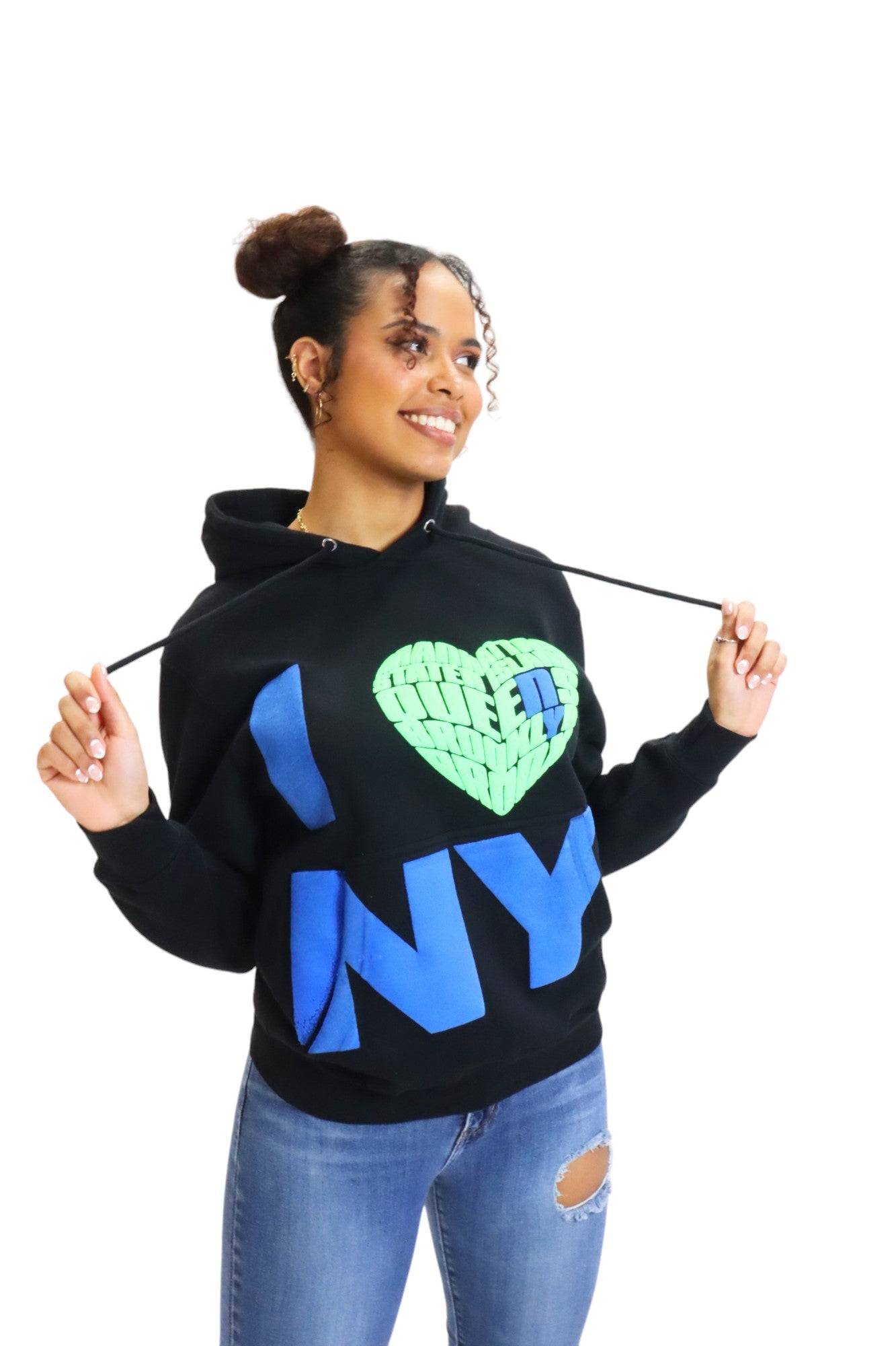 A woman wearing a black hoodie pulls on the drawstrings, showcasing the 3D puff-print 'I ❤️ NY' graphic with its textured green, brick-patterned heart.