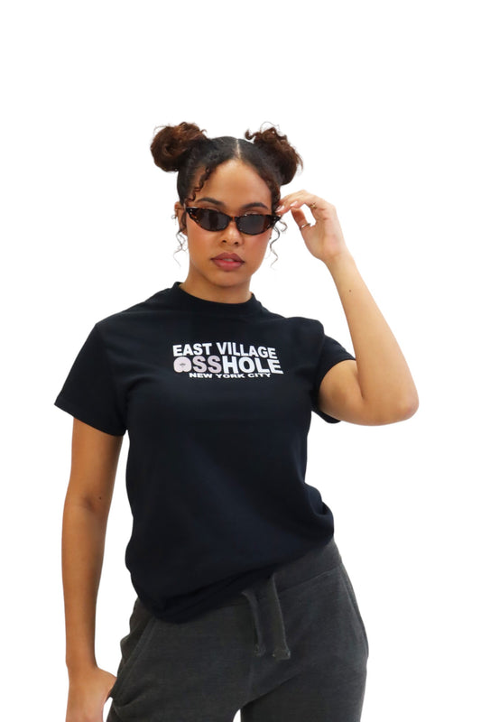 A woman models a black t-shirt with the words 'EAST VILLAGE ASSHOLE' on the chest. The 'O' in 'ASSHOLE' is replaced with a pink graphic of a butt.