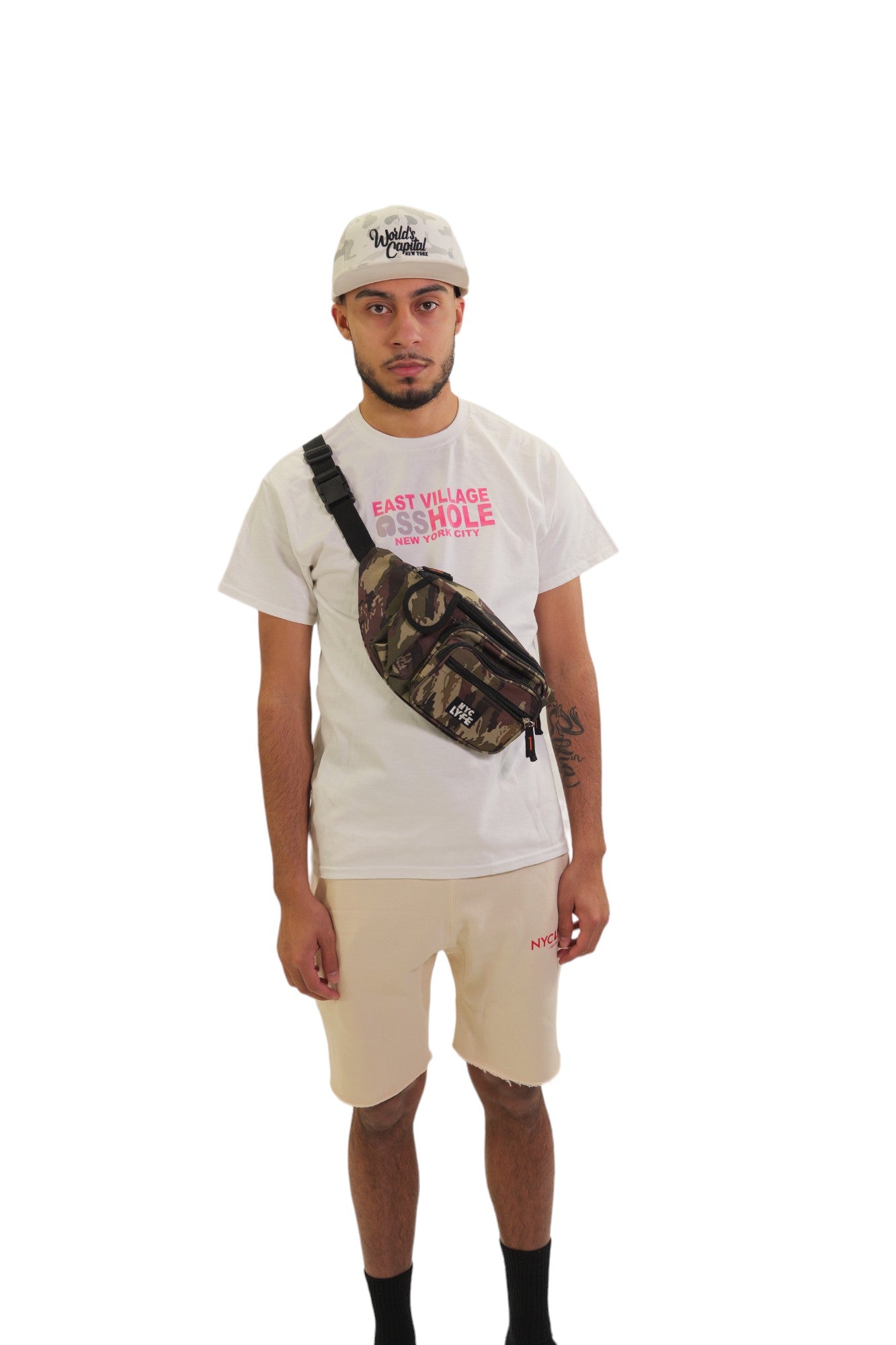 A model stands wearing a full NYC Lyfe outfit, including a white 'East Village Asshole' t-shirt, off-white fleece shorts, a white camouflage 'World's Capital' hat, and a woodland camouflage sling bag across his chest.
