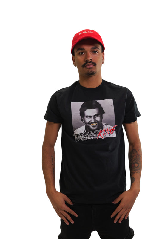 A young man with a mustache poses in a black short-sleeve t-shirt and a red baseball cap against a white background. The t-shirt has a graphic of Pablo Escobar with his teeth colored gold and bold red text over it that says 'Trafficking King'. The red cap has 'Hustle Mentality' embroidered in white.