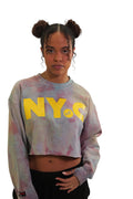 A woman models a long-sleeve cropped crewneck with a grey and pink storm-dye pattern. The letters 'NYC' are printed across the chest in large, bold yellow letters, with a small 'MF' logo inside the letter C.