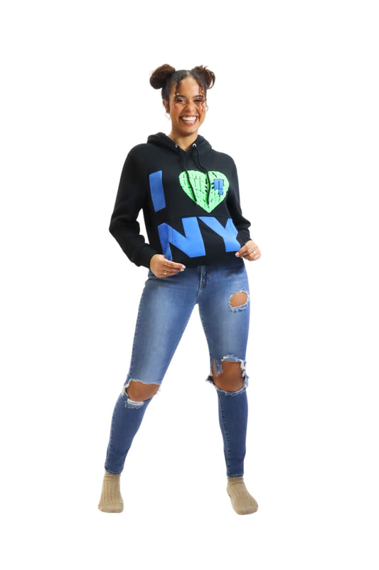A woman smiles while modeling a black hoodie. The front has a large 'I ❤️ NY' graphic with blue letters and a textured, bright green heart.